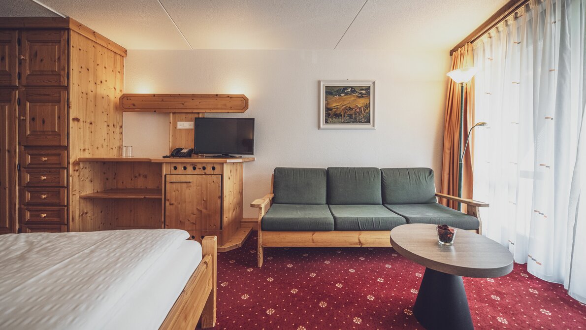 Doppelbettzimmer mit rotem Teppichboden, Fenster und Vorhängen, Sofa mit Tischchen, Inneninterieur aus Arvenholz | © Davos Klosters Mountains 
